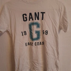 Vit GANT t-shirt med tryck - Vit t-shirt från GANT med stort tryck på framsidan: 'GANT', '1949', 'East Coast' och en blågrön G. Klassisk rund hals och korta ärmar. Materialet är mjuk bomull, perfekt för en chill och sportig look.