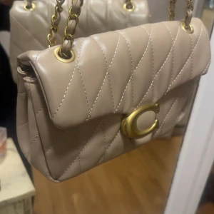 Beige quiltad axelväska från Coach - Säljer en beige quiltad axelväska från Coach med guldfärgad kedjerem och ikoniskt C-spänne framtill. Väskan är tillverkad i mjukt skinn och har ett elegant rutmönster. Perfekt storlek för det viktigaste och ger en lyxig touch till din outfit. Det är inte äkta kommer med dustbag 