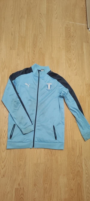 Ljusblå Malmö FF Puma träningsjacka - Snygg ljusblå träningsjacka från Puma med Malmö FF-logga på bröstet. Jackan har mörkblå detaljer längs axlar och ärmar, dragkedja framtill och två fickor med dragkedja. Materialet är mjukt och syntetiskt, perfekt för sport och fotboll.. har några fläckar som syns i mitten av tröjan annars inget fel på den 