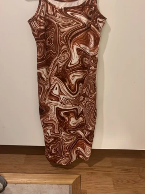 Brun och vit mönstrad ärmlös klänning - Säljer en ärmlös klänning med coolt virvlande mönster i brunt och vitt. Klänningen har en rak, kroppsnära passform och är knälång. Perfekt för dig som gillar unika prints och vill sticka ut med en modern look.