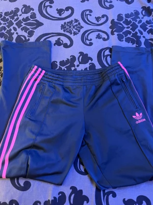 Adidas blå träningsbyxor med rosa ränder - Snygga grå träningsbyxor från Adidas med klassiska rosa ränder längs sidorna och Adidas-logga på benet. Byxorna har elastisk midja med snörning och dragkedjeförsedda fickor. Perfekta för chill eller sport. #y2k #swag #2000s de är mer åt det grå hållet än det blå
