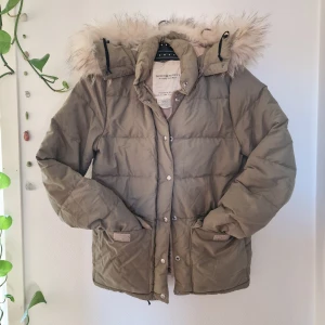 Khakifärgad pufferjacka från Ralph Lauren - Olivgrön/khaki pufferjacka som är stoppad med dun, och med fluffig fuskpäls på huvan (avtagbar), dragkedja och knappar framtill samt 2 fickor fram samt 1 unnerficka. Dragsko i midjan samt i nederkant. 