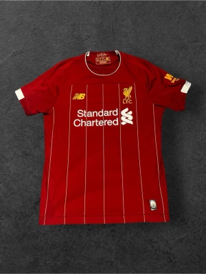 Liverpool matchtröja 2019-2020 - Bilderna visar en röd Liverpool FC matchtröja från New Balance med vita ränder och detaljer, samt Bob Paisley-signatur i nacken. Ingen sko syns på bilderna, så ingen skobeskrivning kan ges.