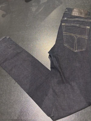 Mörkblå jeans tiger of Sweden 28/32 - Snygga mörkblå jeans från tiger of Sweden med bra  passform och klassiska fem fickor. Jeansen har kontrasterande gula sömmar och ett svart läderpatch bak i midjan. Materialet är slitstarkt denim och modellen sitter tight längs benen. 28/32