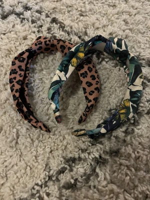 Två snygga hårband med mönster - Säljer två hårband, ett med leopardmönster i brun och svart och ett med blommigt mönster i grönt, gult och vitt. Båda har twistad knut framtill och är tillverkade i mjukt tyg. Typ helt nya och i jättebra skick🫶🏼💕