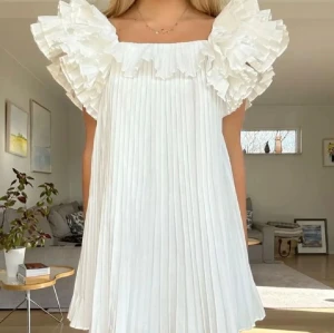 Vit plisserad kortklänning med volang - Säljer denna så fina vita kortklänning med plisserat tyg och stora volangdetaljer vid axlarna. Klänningen har en loose passform och är ärmlös med fyrkantig urringning. Perfekt för dig som gillar romantisk och trendig stil❤️!! Sista bilden är min egen