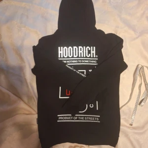 Svart HOODRICH hoodie med tryck - Svart hoodie från HOODRICH med stor vit logga och tryck både fram och bak. Tröjan har huva med dragsko och känguruficka. Snygga grafiska detaljer i vitt och rött ger streetkänsla. Perfekt för dig som gillar urban stil.