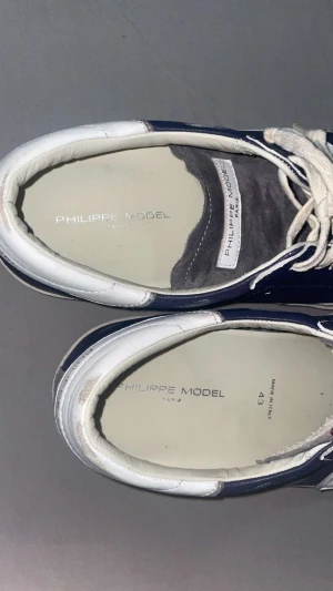 Philippe Model blåvita sneakers - Snygga sneakers från Philippe Model i marinblått och vitt med grå mockadetaljer på tån. Skorna har snörning, rund tå och platt sula med tydlig logga under. Tillverkade i skinn och mocka, klassisk låg modell med vita kontraster och stilren design.