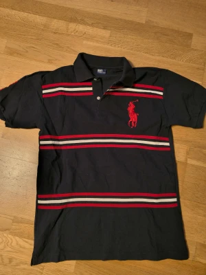 Svart randig polotopp från Polo Ralph Lauren - Svart polotopp med röda och vita ränder från Polo Ralph Lauren. Stor röd broderad logga på bröstet, klassisk krage och två knappar framtill. Tillverkad i mjuk bomull och har korta ärmar. Snygg och sportig stil med tydliga kontraster.