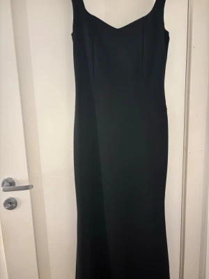 Svart långklänning med v-ringning - Stilren svart långklänning med v-ringad hals och breda axelband. Klänningen har en figurnära passform och är helt slät utan mönster eller detaljer. Perfekt för dig som gillar minimalistisk och elegant stil.