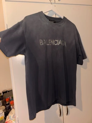 Mörkgrå Balenciaga t-shirt med paljetter - Snygg mörkgrå t-shirt från Balenciaga i unisex-modell, storlek S. Framsidan har Balenciaga-loggan i silvriga paljetter och baksidan har ett stort BB-emblem. T-shirten är i mjuk bomull och har en klassisk rund hals och korta ärmar.