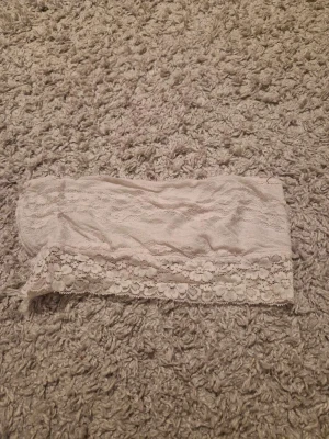 Ljusbeige spets bandeau från New Woman - Supersöt bandeau tubtopp i ljusbeige från New Woman. Toppen är ärmlös och har ett vackert spetslager med blommönster längs nederkanten. Materialet är mjukt och stretchigt, perfekt för att styla med höga jeans eller kjol. Spetsen ger en romantisk och feminin vibe.