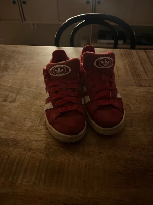 Röda Adidas sneakers med vita ränder - Säljer ett par röda Adidas sneakers med klassiska vita ränder på sidorna och Adidas logga på plösen och hälen. Skorna har snörning och en platt vit sula. Ovandelen är i mocka och de har en sportig, retro vibe som passar perfekt till streetwear.