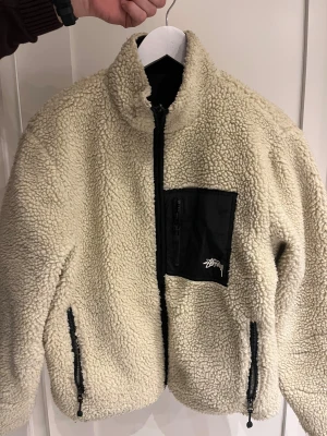 Stüssy teddyjacka med 8-ball - Säljer en teddyjacka från Stüssy i beige och svart med stor 8-ball på ryggen. Jackan är vändbar med ena sidan i fluffig teddy och andra i svart tyg med Stüssy-logga. Flera dragkedjor och cool bröstficka. Perfekt för dig som vill sticka ut.
