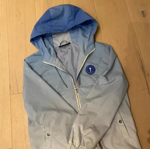 Trapstar vindjacka-blå/vit - Snygg blå vindjacka från Trapstar med huva och dragkedja med unikt T-formad zip. Jackan har broderad Trapstar-logga på ryggen och rund badge framtill. Ljusblå och mörkblå färgblock, elastiska ärmslut och praktiska fickor. Perfekt för dig som vill sticka ut med en jävligt fräsch jacka!
