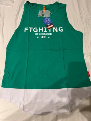 Grön ärmlös linnetopp med RUN-tryck - Säljer en grön ärmlös linnetopp från KFIARXOUR med vit text 'FTGHITNG' och 'RUN' tryckt på fram- och baksidan. Toppen har en vit kant längst ner och rund hals. Perfekt för dig som gillar sportig stil och vill sticka ut på träningen.