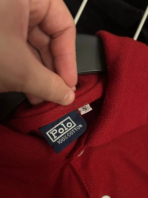 Röd Polo pikétröja med svart logga - Snygg röd pikétröja från Polo i 100% bomull. Klassisk krage, två vita knappar och stor svart broderad logga på bröstet. Kortärmad och perfekt för en chill och stilren look. Passar dig som gillar sportig vibe och enkel design.