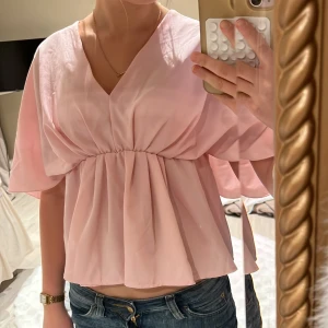 Ljusrosa blus från Meet Me There - Säljer en ljusrosa blus från Meet Me There i storlek XXS/XS. Blusen har v-ringad hals, breda korta ärmar och ett snyggt rynkat midjeparti som ger en flowy och chill siluett. Perfekt för dig som gillar mjuka pasteller och en feminin look.