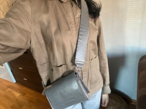 Grå axelväska med croco-mönster från cw<3 - Snygg grå axelväska med croco-präglat mönster och bred justerbar rem. Väskan har en stilren, rektangulär form och är tillverkad i skinnimitation. Metallspänne på remmen för enkel justering❤️ord pris 499kr