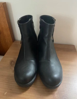 Svarta boots med klack och spänne - Snygga svarta boots i skinn med rund tå och låg klack. Dragkedja på sidan och dekorativt spänne i metall ger en cool vibe. Perfekta till jeans eller kjol när du vill ha en stilren look. Platt sula och robust design för extra komfort.