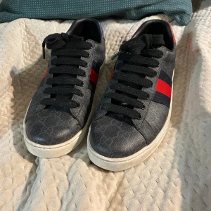 Gucci sneakers med röd/blå rand - Snygga svarta Gucci sneakers med klassisk röd och blå rand på sidan och diskret GG-mönster över hela skon. Skorna har svarta snören och vit sula, tillverkade i skinn med textildetaljer. Perfekta för dig som vill ha en lyxig och trendig look.