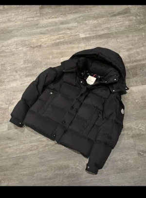 Svart dunjacka från Moncler - Säljer en svart dunjacka från Moncler med huva och klassisk logga på ärmen. Jackan är tjockt vadderad, har dragkedja och knappar framtill samt praktiska fickor. Perfekt för kalla dagar och riktigt snygg streetstil.