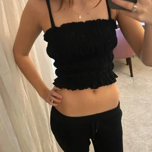 Svart croppad linnetopp med volang - Säljer en svart croppad linnetopp med smala axelband och volangkant nedtill. Toppen har en smockad, ribbad struktur som ger en snygg och figurnära passform. Perfekt för dig som gillar en enkel men trendig look.