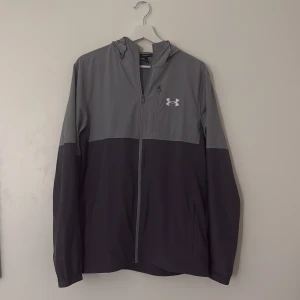 Grå och svart vindjacka Under Armour - Snygg tvåfärgad vindjacka från Under Armour i grått och svart. Jackan har huva, hel dragkedja framtill och diskret logga på bröstet. Tillverkad i lätt syntetmaterial, perfekt för aktiva dagar. Långärmad och med sportig vibe. Den säljs inte längre på nätet och den har blivit sällsyn. Bara att skriva ifall man vill ha fler bilder och pris kan diskuteras vid snabbaffär.