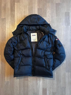 Svart pufferjacka från Hilfiger Denim - Snygg svart pufferjacka från Tommy Hilfiger Denim med huva och dragkedja. Perfekt för kalla dagar och riktigt skön att bära ❄️. Det står L men passar mer som en M, personen på bilden är 183. Skick 9.5/10 ✅Nypris cirka 2700