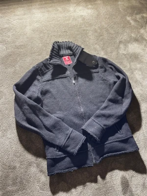 Vintage J&J cardigan👖 - Vintage Cardigan från Jack & Jones med hög ribbad krage och dragkedja framtill. Jackan har långa ärmar och en snygg detalj med knapp vid halsen. Perfekt för dig som gillar en clean och stilren look.