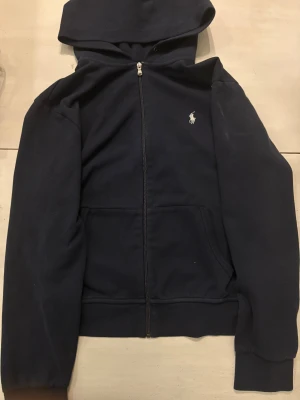 Mörkblå hoodie från Polo Ralph Lauren - Snygg mörkblå hoodie från Polo Ralph. Väldigt skön men har dock några små tecken på användning. En på sista bilden att texten har försvunnit och vid för man spänner åt den att det inte finns något snörre den gick sönder och tog bort den. Nypris 2000kr