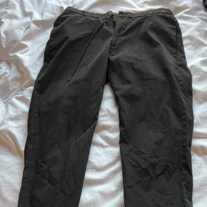 Svarta tapered byxor med snörning - Snygga svarta byxor med tapered passform och elastisk midja med snörning. Byxorna har diskreta fickor både fram och bak, samt en liten metallring vid bakfickan. Perfekta för en chill och stilren look.