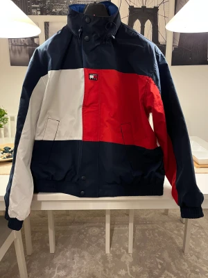 Kith x Tommy Hilfiger Reversible vindjacka - Jacka i färgerna marinblå, vit och röd. Jackan har en avtagbar huva och stängs med knappar och dragkedja. På ryggen står det "TOMMY KITH". Framsidan har en liten Tommy Hilfiger logga. Reversible. Helt ny utan prislapp.