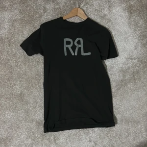 Svart RRL t-shirt från Ralph Lauren - Svart t-shirt från Ralph Lauren Double RL med stor grå RRL-logga på bröstet. Klassisk rund hals och korta ärmar. Tillverkad i mjuk bomull som känns skön mot huden. Perfekt för dig som gillar stilrena och ikoniska tryck. Passform slimfit