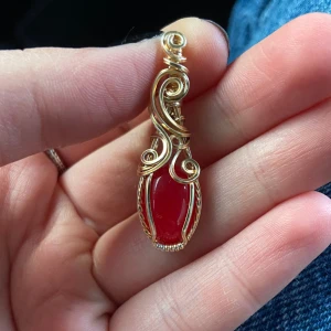 Handgjord guldberlock med röd sten - Unik berlock med en intensivt röd, oval sten infattad i snirklig guldfärgad metalltråd. Hängsmycket har vackra detaljer och en elegant, bohemisk känsla. Perfekt för dig som vill sticka ut med ett personligt smycke.