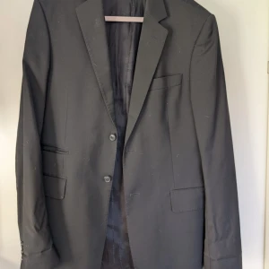 Svart kavaj från Tiger of Sweden - Stilren svart kavaj från Tiger of Sweden med klassisk tvåknappsdesign och snygga fickdetaljer. Kavajen har slits baktill och är helfodrad med diskret mönstrat tyg. Passformen är normal och materialet känns mjukt och följsamt, perfekt för dig som gillar en clean look.