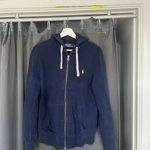 Marinblå hoodie från Polo Ralph Lauren - Snygg marinblå hoodie från Polo Ralph Lauren med gul broderad logga på bröstet. Tröjan har vit snörning i huvan, dragkedja framtill och två stora fickor. Perfekt för chill dagar och har en klassisk streetstil.