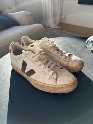 Vita Veja sneakers med grön detalj - Säljer ett par klassiska Veja sneakers i vitt med grå V-logga på sidan och grå hälflik med VEJA-tryck. Skorna har snörning, platt sula och är tillverkade i skinn och textil. Perfekta för dig som gillar stilrena och hållbara sneakers.