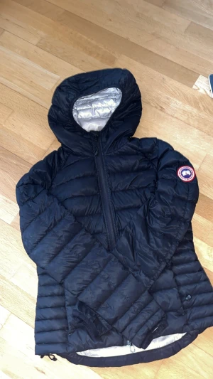Svart dunjacka från Canada Goose - Snygg svart dunjacka från Canada Goose med silvrigt foder och huva. Jackan har ett quiltat mönster, dragkedja framtill och klassisk logga på ärmen. Perfekt för kalla dagar och har en clean, sportig look som funkar till det mesta.ÄKTA