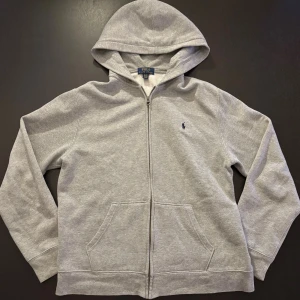 🩶 Grå zip-hoodie från Polo Ralph Lauren – Storlek Small - Säljer min mysiga och stilrena hoodie från Polo Ralph Lauren 👕✨. Den är i en klassisk ljusgrå färg som funkar till allt och har en bekväm huva 🧢 samt dragkedja hela vägen 🔛. Materialet är mjukt, varmt och perfekt för både vardag och träning 💪🔥.  Den broderade loggan på bröstet ger tröjan en snygg och tidlös look 🐎💙. Passformen är en normal Small och sitter riktigt bra utan att vara för tight 🙌.  Det här är verkligen en hoodie som passar i alla lägen. Först till kvarn.