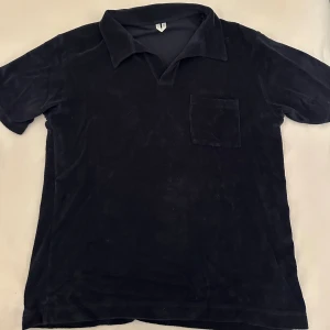 Mörkblå frottépiké från Arket - Mjuk och chill mörkblå piké från Arket i frotté, med klassisk krage och en bröstficka. Skjortan har korta ärmar och loose passform, perfekt för en avslappnad stil. Tillverkad i Portugal och känns riktigt skön mot huden.