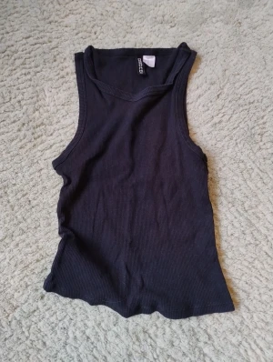 Svart ribbad linnetopp från H&M Divided - Svart ribbad linnetopp från H&M Divided i storlek XS. Toppen är ärmlös med rundad hals och har en slim passform som sitter snyggt mot kroppen. Perfekt att styla med jeans eller kjol för en enkel och cool look.