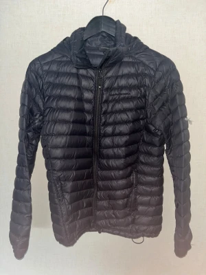 Svart dunjacka Peak Performance - Svart dunjacka från Peak Performance med huva och dragkedja framtill. Jackan är quiltad och har en clean, sportig look. Perfekt för kyliga dagar och enkel att styla till det mesta. Snygg passform och diskret logga på bröstet. Jackan har synliga repor inne i jackan därför de låga priset.