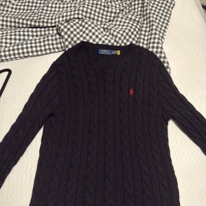 Svart kabelstickad tröja Polo Ralph Lauren - Svart kabelstickad tröja från Polo Ralph Lauren med klassisk rund hals och röd broderad logga på bröstet. Tröjan är tillverkad i mjuk bomull och har långärmad passform med ribbade muddar vid ärmslut och nederkant. Perfekt för en clean och stilren look. Som ni ser, köpt från Care of Carl den 27 januari 2025 för 2199 kr. Den är ny och bara använd 4 gånger, finns inga tecken på användning eller någon slitning.