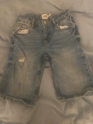 Blå denimshorts från 157, storlek 140 - Säljer ett par klassiska blå jeansshorts från 157 i storlek 140. De har fem fickor, slitna detaljer och råa kanter vid bensluten för en chill vibe. Materialet är robust denim med knapp och dragkedja framtill. Perfekt för dig som gillar streetstyle.