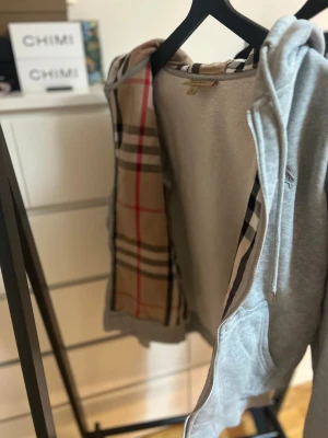 Grå Burberry hoodie med dragkedja - Snygg grå hoodie från Burberry i storlek M med klassiskt rutigt foder i huvan. Tröjan har dragkedja, snörning vid huvan och två fickor framtill. Ikonisk Burberry-logga broderad på bröstet. Perfekt för dig som gillar stilrena och lyxiga streetwear-plagg. Tags finns pm vid bild!