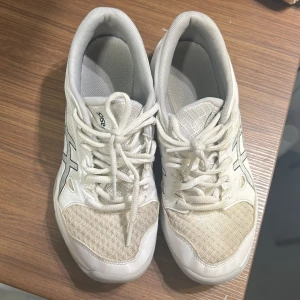 Vita Asics Gel-Rocket 11 sneakers - Säljer ett par vita Asics Gel-Rocket 11 sneakers i storlek 37. Skorna har mesh och syntet på ovansidan, med klassisk Asics-logga i svart på sidan. Platt sula i grått och vitt, snörning framtill och rund tå. Perfekta för dig som gillar sportig stil.