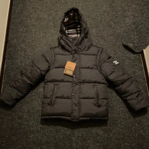 Svart dunväst Burberry jacka med huva - Säljer en dunväst burberry jacka med avtagbara ärmar och huva. Västen har två fickor med dragkedja och klassiskt rutigt foder i svart, vitt och rött. Stängs med både dragkedja och knappar framtill. Perfekt för kalla dagar när du vill ha en clean look. Skriv om du är intresserad. Pris kan diskuteras!!