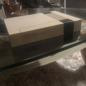 Nintendo Entertainment System (NES) med spel och kontroller - Klassisk NES-konsol i gott skick med två originalkontroller och tre populära spel: Super Mario Bros, Dr. Mario och Super Mario Bros 3. Konsolen och tillbehör visar lättare bruksspår men är fullt funktionella. Perfekt för retrospelare och samlare!