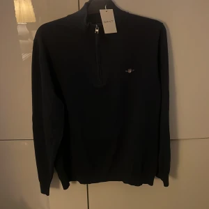 Mörkblå half zip tröja från GANT - Snygg mörkblå tröja från GANT i bomull med half zip-dragkedja och diskret logga på bröstet. Klassisk och stilren design med långärmade ärmar och mjuk känsla. Perfekt för dig som gillar en clean look och vill ha något som funkar året runt.